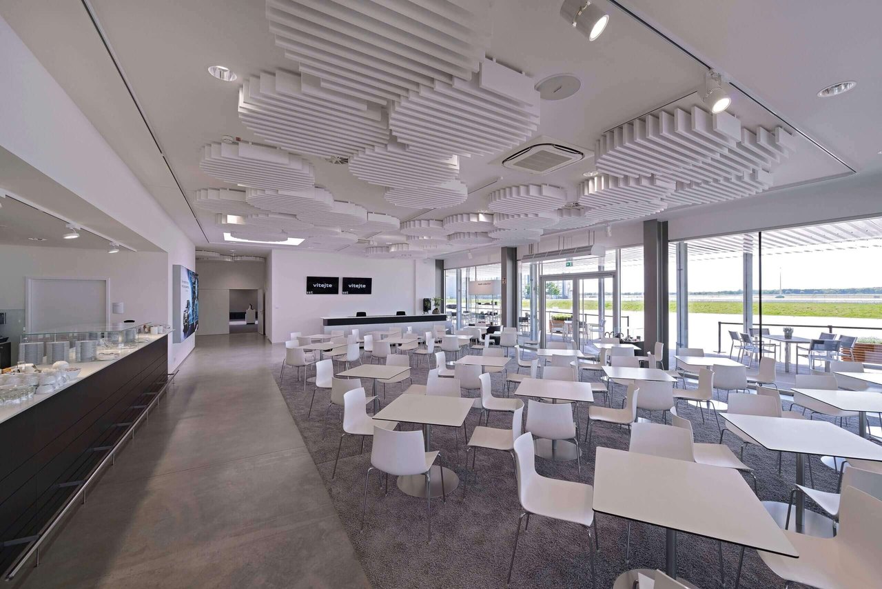 Heller Konferenzraum mit weißen Tischen und Stühlen, kreisförmigen Deckenpaneelen und Blick auf einen Flughafen.