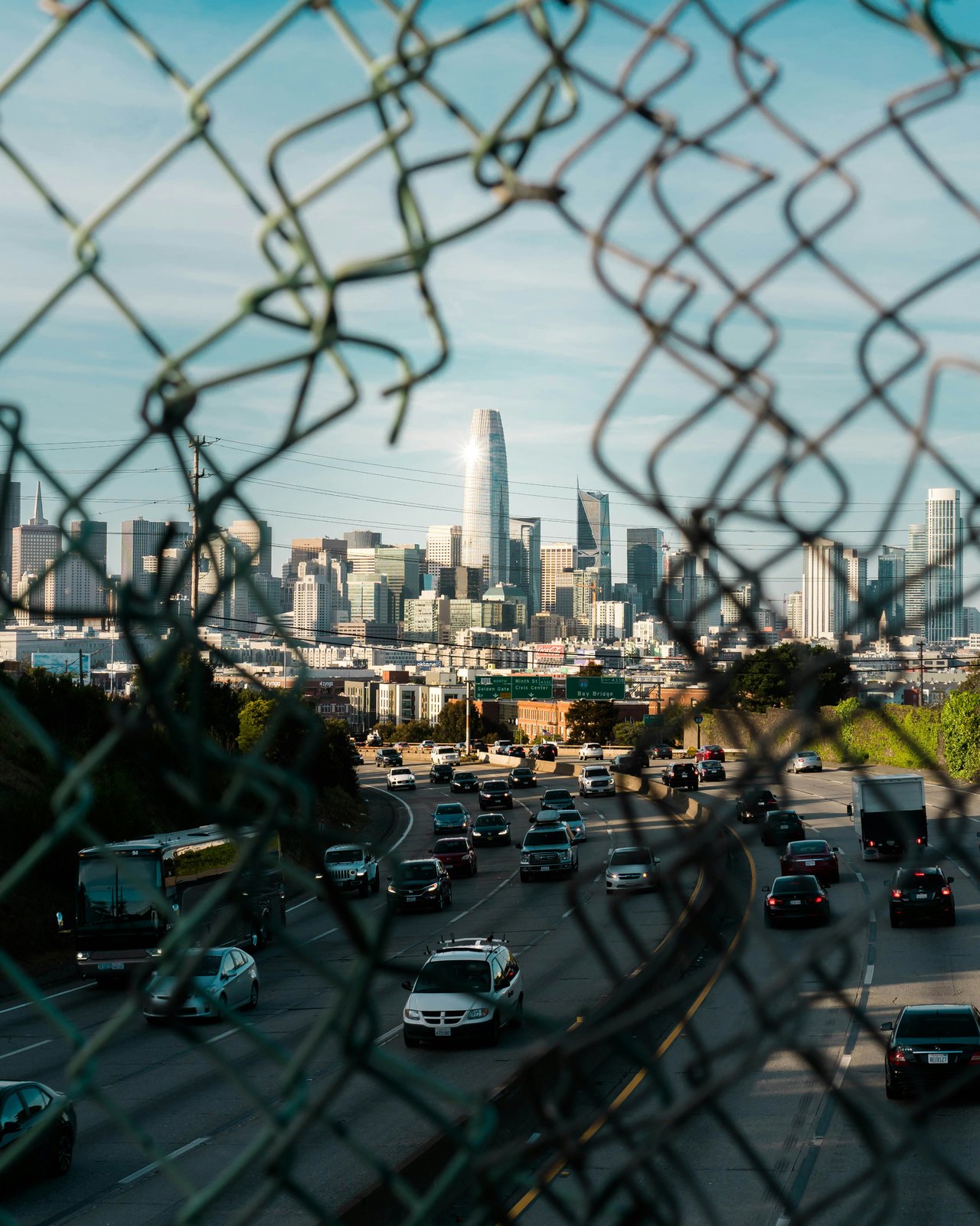 Blick durch einen Maschendrahtzaun auf die Skyline von San Francisco und eine belebte Autobahn bei Tageslicht.