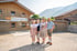 Familie in Tracht steht vor einem Holzhaus mit dem Schild „Herzlich Willkommen“, Berge im Hintergrund.