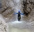 Eine Person in Taucheranzug und Helm steht unter einem Wasserfall in einer Felsenschlucht.
