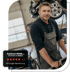 Ein Mechaniker in dunkelblauem Hemd und grauer Mazda-Latzhose in einer Autowerkstatt mit angehobenen Autos im Hintergrund.