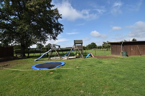 Spielplatz im Freien mit Schaukeln, Rutschen, Sandkasten, Trampolin und Baum unter blauem Himmel mit Wolken.