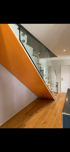 Moderne Treppe mit orangefarbener Wand, Holzstufen und Glasgeländer in einem hellen Raum.