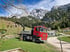 Roter LKW mit Kranarm auf Schotterweg in den Alpen vor schneebedeckten Bergen.