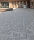 Grey rectangular paving stones in a herringbone pattern cover a wide outdoor plaza leading to a modern wooden building.