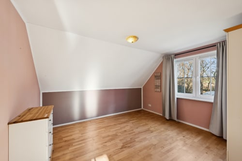 Leeres Schlafzimmer mit Dachschräge, holzfarbenem Boden, rosa und braunen Wänden, Fenster mit Gardinen und Kommode.