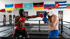 Zwei Boxer in Kopfschutz und Handschuhen kämpfen in einem Ring; Flaggen im Hintergrund.