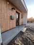 Moderner Hauseingang mit Holzfassade, schwarzer Haustür, Briefkasten, Hausnummer 5 und grauem Betonsockel.