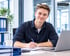 Ein lächelnder junger Mann im dunklen Hemd sitzt am Schreibtisch mit Laptop und schreibt in ein Notizbuch.