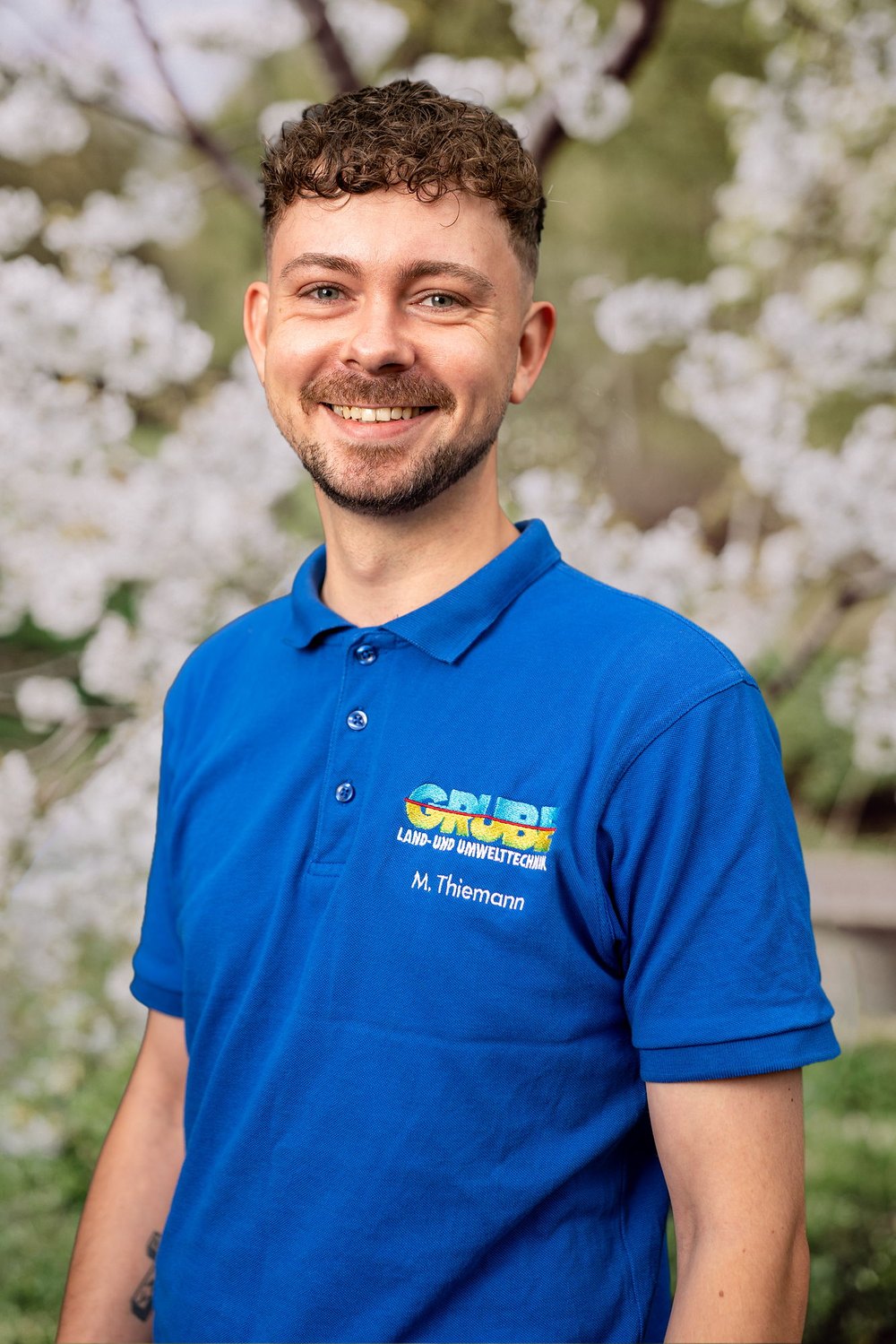 Frau mit grauen Haaren und Brille in blauem Poloshirt mit "GRÜN" Logo vor weiß blühendem Baum.