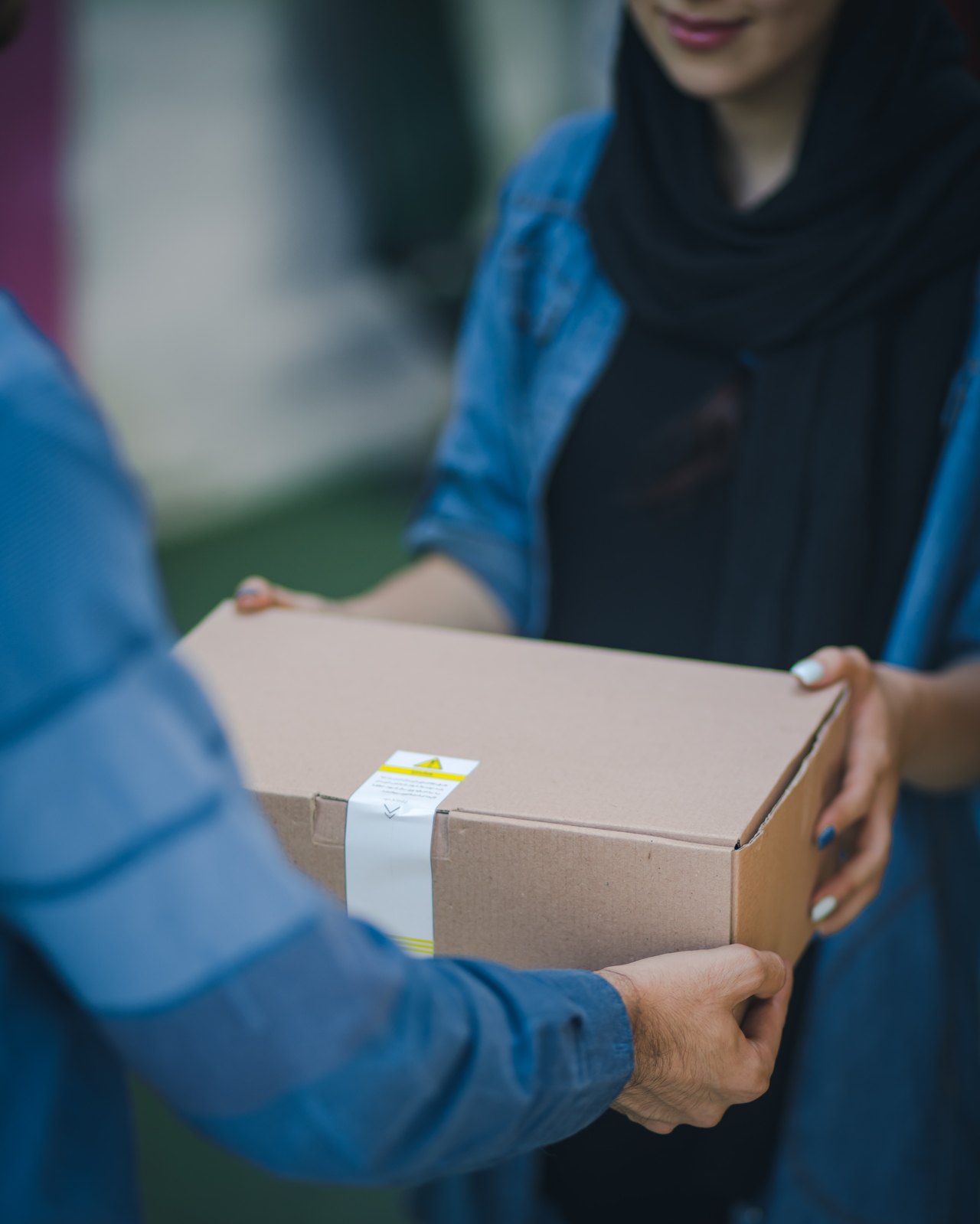 Eine Frau mit schwarzem Hidschab nimmt ein braunes Paket von einem Zusteller in blauer Kleidung entgegen.