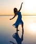 Eine Frau in einem blauen Kleid tanzt barfuß im seichten Wasser bei Sonnenuntergang.