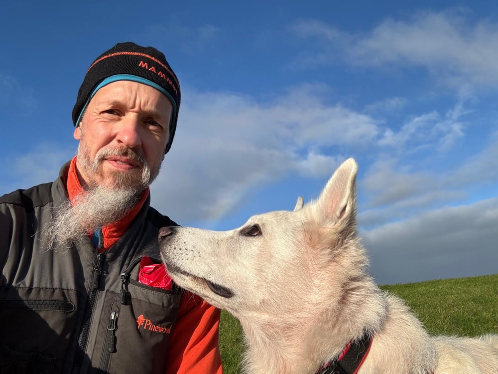 Mann mit Bart und Mütze neben weißem Hund vor blauem Himmel mit Wolken.