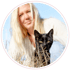 Eine blonde Frau im Hintergrund mit einer schwarzen Katze im Vordergrund, die in Gras sitzt.