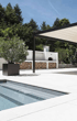 Ein Poolbereich mit einer Pergola, einer weißen Außenküche mit Holzstapeln und einem Olivenbaum in einem Topf.