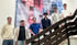 Sechs junge Männer posieren lächelnd auf einer Treppe vor einem großen blauen Werbebanner mit QR-Code und Porträtfoto.