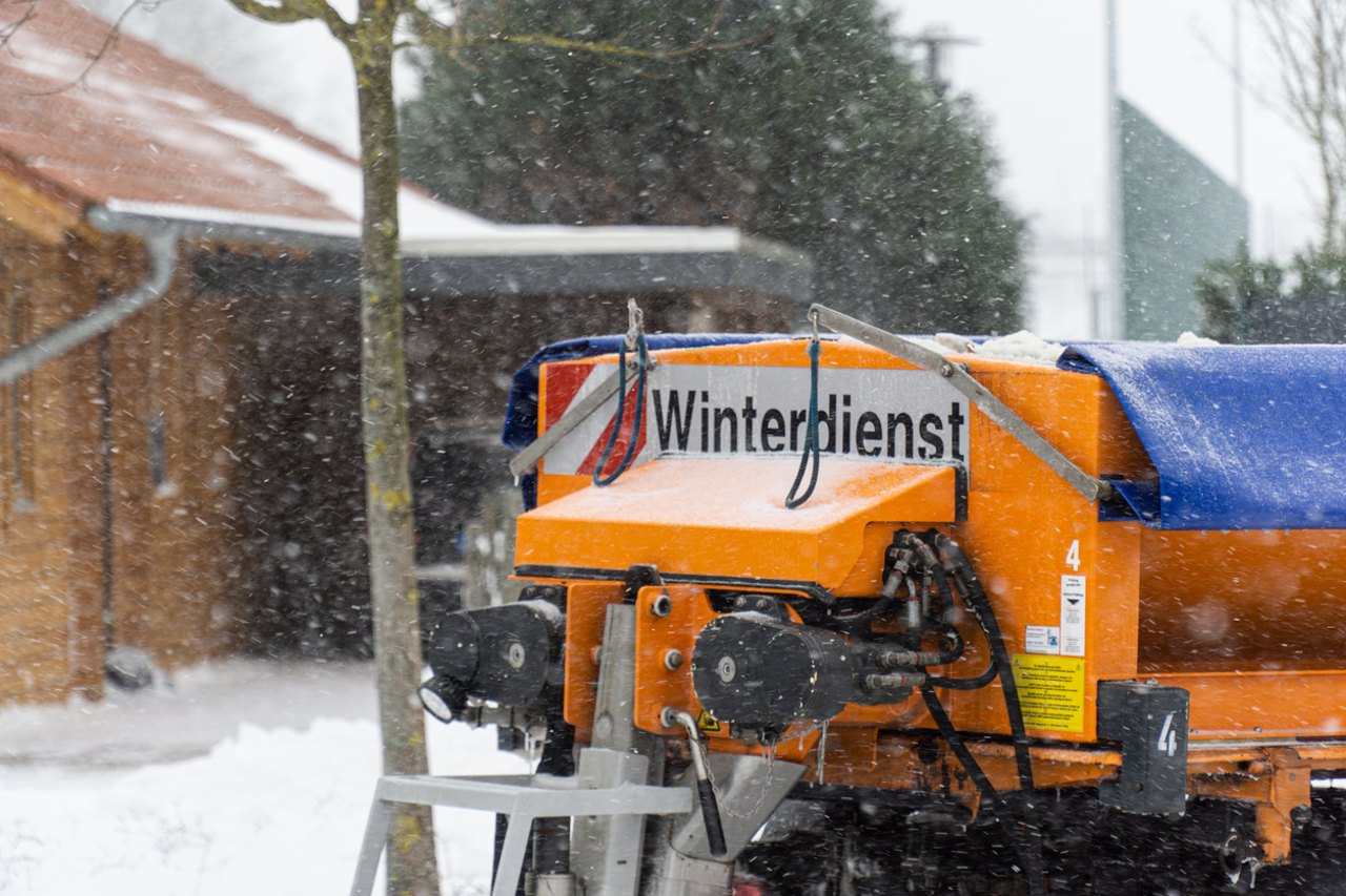Oranger Winterdienst-Streuwagen mit Schnee und Bäumen im Hintergrund.