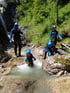 Vier Personen in Neoprenanzügen und blauen Helmen klettern durch einen felsigen Bachlauf mit Wasserfällen.