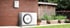 Eine weiße Bosch-Wärmepumpe und ein kleineres Wandgerät an einer roten Backsteinwand neben einem Garten.