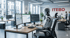 Ein humanoider Roboter sitzt an einem Schreibtisch mit drei Monitoren in einem modernen Büro mit dem Logo "ITEBO".