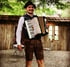 Mann in Lederhosen und Hut spielt Akkordeon vor einer Holzhütte im Freien.