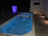 Zwei beleuchtete Whirlpools im Freien bei Nacht mit einer lila leuchtenden Bar und Wandlampen.