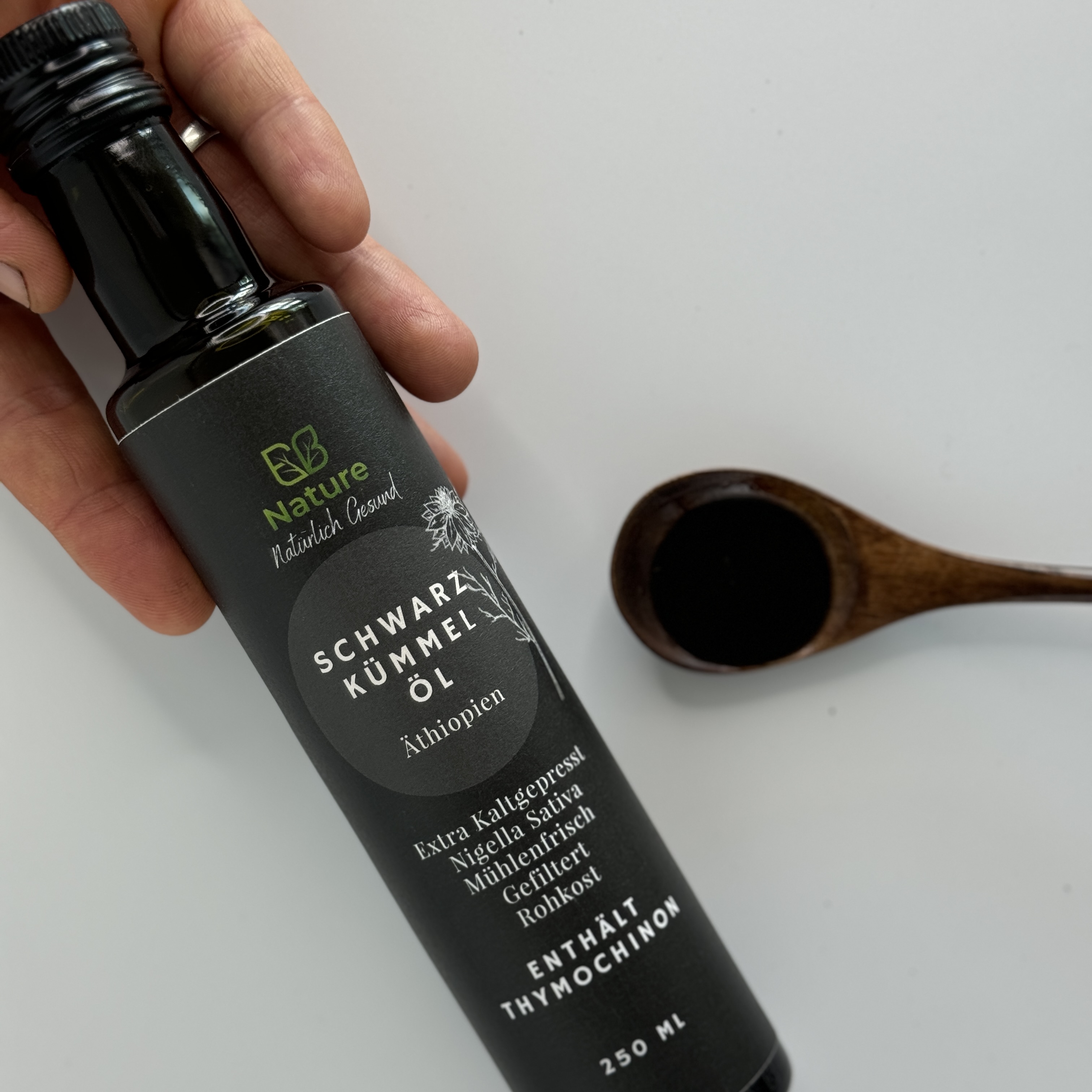 Hand hält EB Nature Schwarzkümmelöl Flasche, daneben Holzlöffel mit dunklem Öl