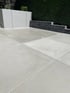 Moderne Terrasse mit großen, hellgrauen Betonplatten, einer weißen Ziermauer und einer dunklen Steinumrandung vor einer Hecke.