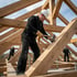 Zimmermann mit Helm montiert Dachbalken auf einem Holzrahmenbau bei Tageslicht