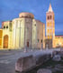 Eine beleuchtete Kirche mit Kuppel und ein hoher Glockenturm bei Dämmerung, umgeben von Steinsäulen und Ruinen,die Stadt nennt sich Zadar.