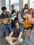 Four children playing electric guitars with an adult sitting on the floor in front of them.