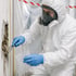 Fachkraft in Schutzanzug und Atemschutz entnimmt eine Materialprobe aus der Wand und füllt sie in einen Probenbeutel