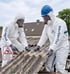 Zwei BauSanTec-Mitarbeiter in weißen Schutzanzügen und Atemschutzmasken entfernen Wellplatten von einem Hausdach.