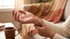 A person holds their wrist with their other hand while sitting indoors near a coffee mug and a blanket.