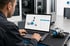 Techniker bedient Laptop mit 3D-Explosionszeichnung eines Bauteils vor einer Industriemaschine in einer Werkshalle.