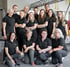 Team von elf Physiotherapeuten in schwarzen Poloshirts posiert in einem Fitnessstudio.
