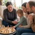 Vier Personen – eine Frau, ein Mann, ein Kind und eine ältere Frau – spielen mit Holzfiguren auf einem Brett.