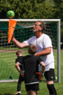 Ein Fußballtrainer hält einen orangefarbenen Kegel mit einem grünen Fußball auf dem Torbalken, vor zwei Jungen.