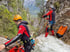 Zwei Personen in Neoprenanzügen und Kletterausrüstung am Rande eines felsigen Wasserfalls.