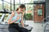 Frau mit Zopf und gelbem Tanktop sitzt auf einer Trainingsbank in einem Fitnessstudio und hält sich mit einer Hand den unteren Rücken