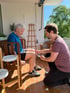 Ein Physiotherapeut kniet vor einem älteren Mann und behandelt dessen Knie auf einem sonnigen Balkon.