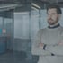 Ein bärtiger Mann im grauen Pullover steht mit verschränkten Armen in einem modernen Büro mit Glaswänden.