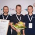 Drei Männer lächeln; einer hält eine Urkunde "Gründungspreis 2024 Finalist", ein anderer einen Blumenstrauß.