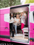 Drei Frauen feiern in einem pinken Mercedes-Benz Lieferwagen, der mit "Powered by Mercedes-Benz" beschriftet ist.