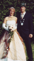 Ein Paar in festlicher Kleidung steht vor einem grünen Busch. Die Frau trägt ein weißes Kleid und hält einen Blumenstrauß.