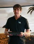 Ein Mann in einem schwarzen Poloshirt mit "VA"-Logo hält ein Tablet vor einem Strandhintergrund.