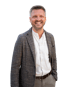 Ein lächelnder Mann mit grauem Haar und Bart trägt einen karierten Blazer über einem weißen Hemd und einer grauen Hose.