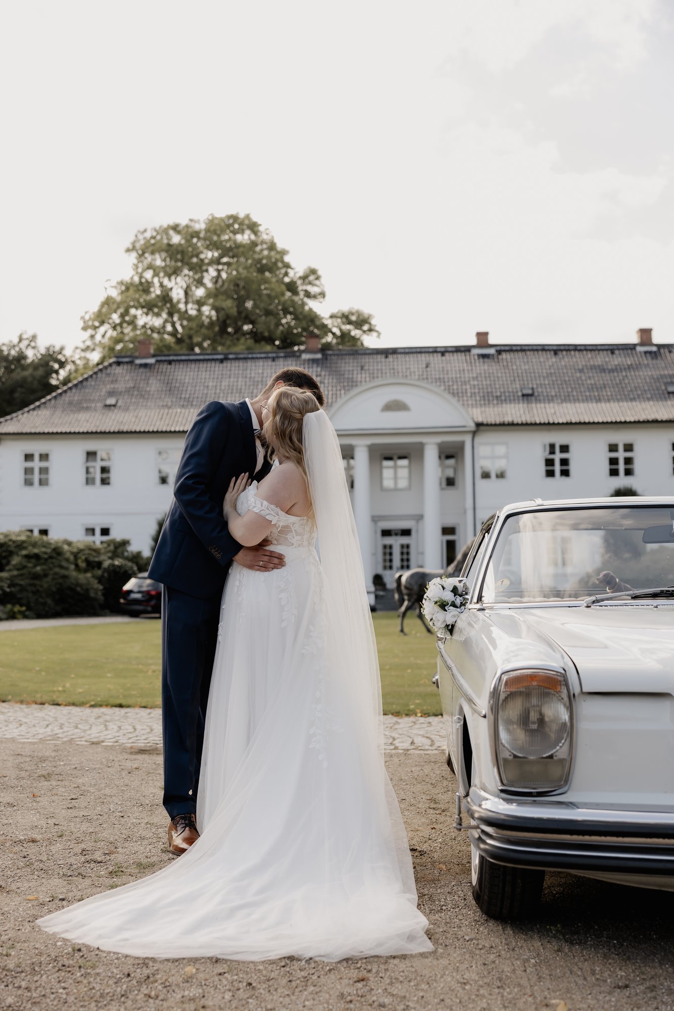 Brautpaar umarmt sich vor einem Oldtimer und einem Gutshaus
