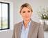 Porträt einer Frau mit blonden Haaren in einem beigen Blazer und hellblauem Hemd vor hellem Hintergrund mit Blumen.