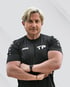 Ein Mann mit blonden Haaren und schwarzem T-Shirt mit "JAKO" und "TP" Logo verschränkt die Arme.